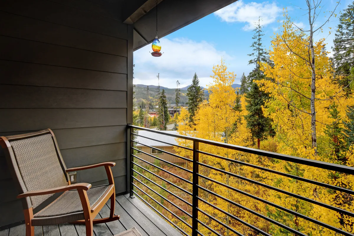 A peaceful balcony with a rocking chair, a playful hanging ornament, and yellow-leaved trees in the background. The scene suggests a connection to the outdoors, ideal for quiet moments, reading, or simply soaking in the surroundings as the seasons change.