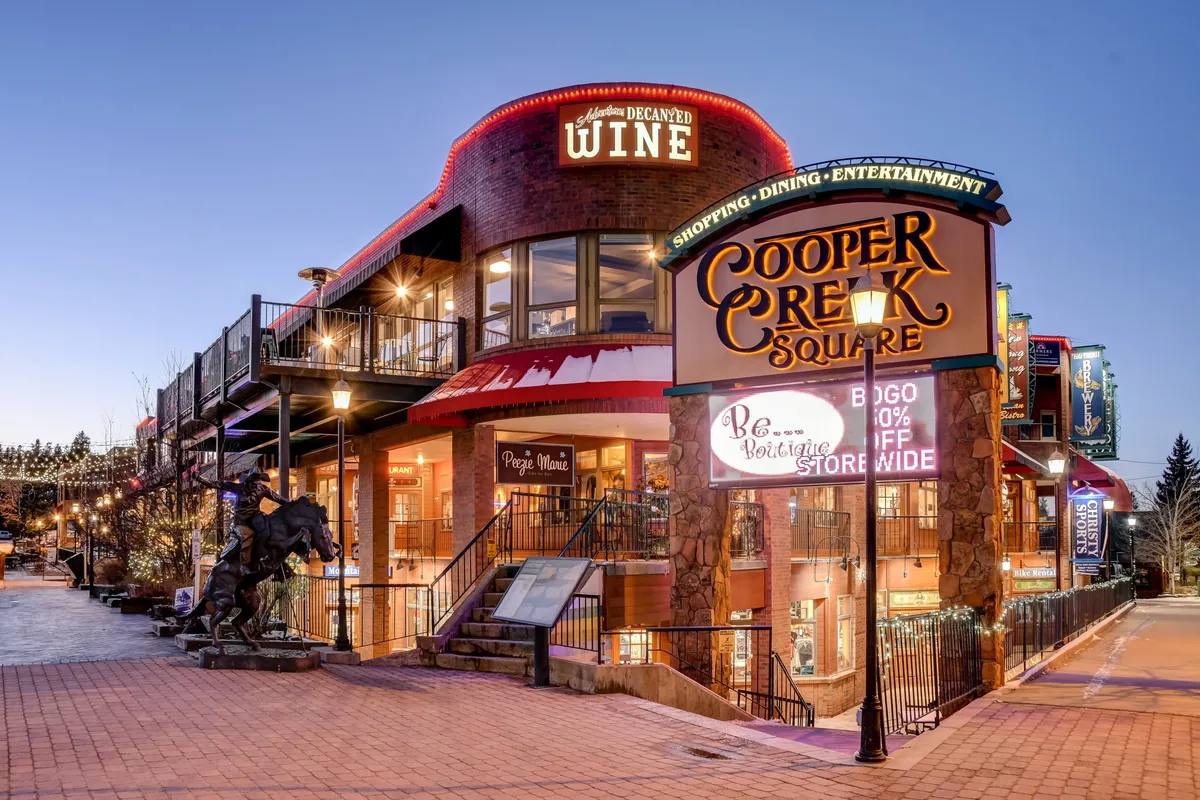 The image shows the vibrant exterior of Cooper Creek Square, featuring its signature sign, inviting outdoor seating, decorative wine signage, a staircase to upper levels, and an artistic statue. The area is lively, evoking a festive mood with elements like live music events and seasonal festivals.