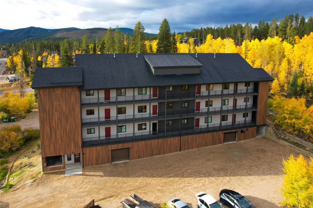 A welcoming modern building facade with colorful window shutters, set against a picturesque landscape of trees and mountains glowing with fall colors. The scene suggests easy access to nature trails and outdoor adventure, with a sense of serenity and possibility.