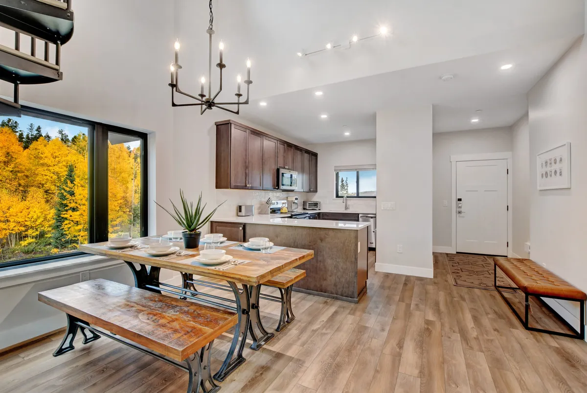 A cozy open-concept space with a dining table set for a meal, a modern kitchen with stainless appliances and dark cabinetry in the background, a large window revealing autumn trees, and a striking spiral staircase connecting the space. Natural light fills the room, emphasizing the seamless flow between kitchen, dining, and living areas.