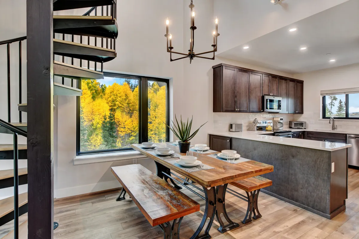 A bright, open interior space featuring a spiral staircase, a large dining table perfect for gatherings, and a generous window offering scenic views. The warm lighting and inviting layout create an ideal central hub for families or groups to socialize and plan their day.