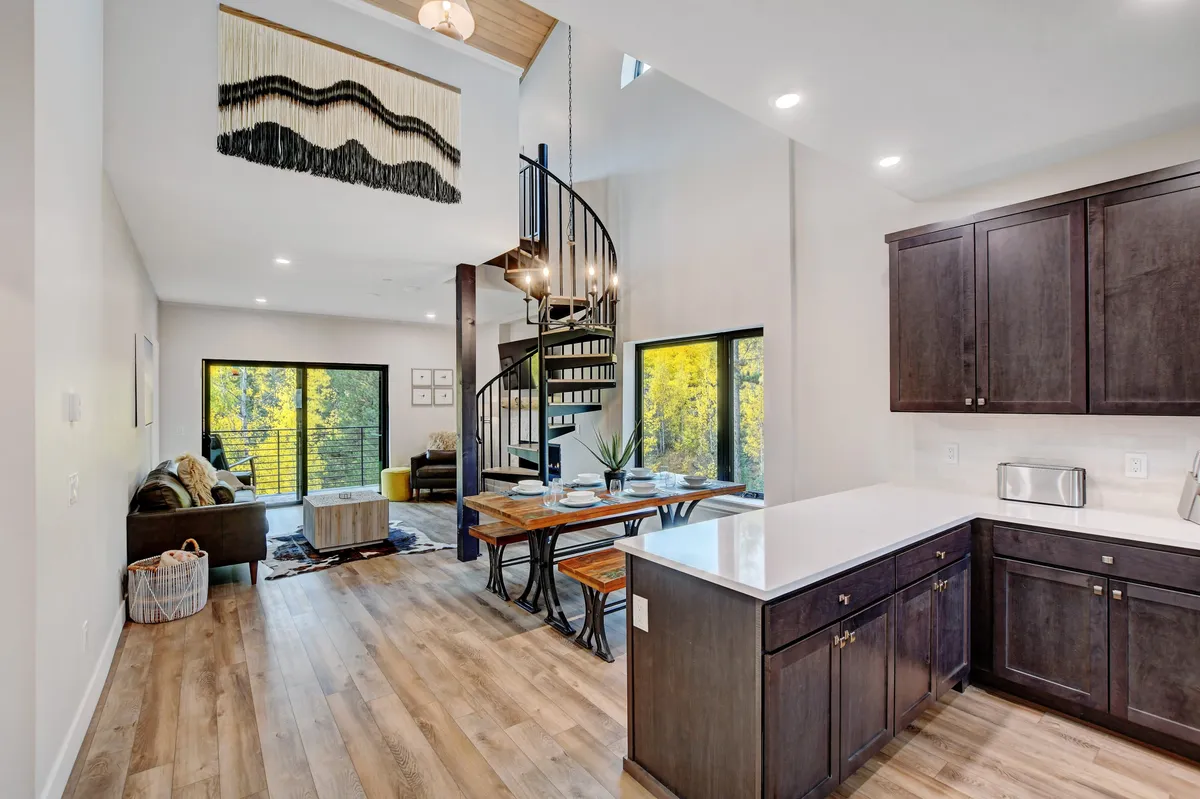 This image shows a welcoming, open-plan dining and living area featuring a spiral staircase, a large family dining table, and expansive windows letting in natural light. The space is contemporary yet cozy, with decorative wall art and thoughtful details that make it suitable for families and groups.