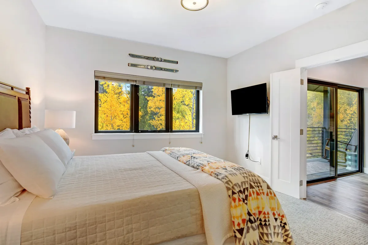 A cozy bedroom with a bed positioned near large windows, decorative skis on the wall, a smart TV, and a door to the balcony. The combination of natural light, decor, and tech amenities makes this room inviting for relaxation or entertainment.