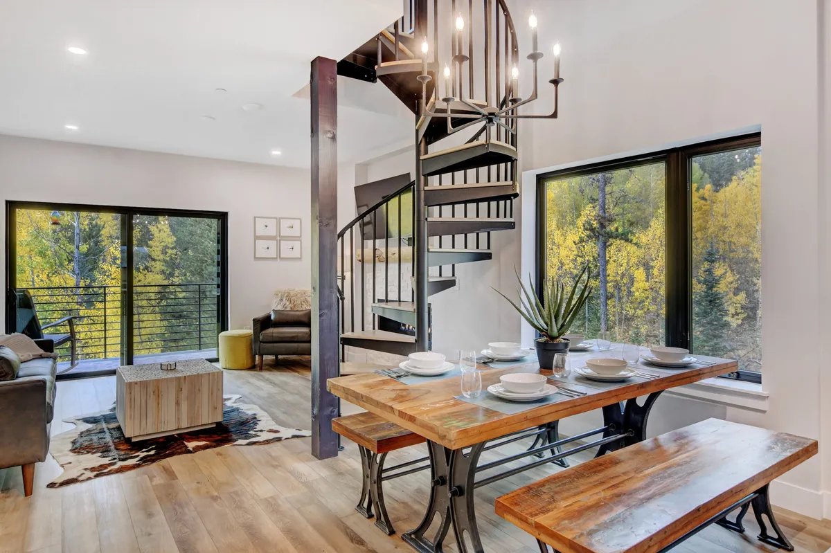 This image shows a spacious interior highlighted by a dramatic spiral staircase leading up to a lofted area. The cozy living space benefits from large windows that fill the room with natural light, and a dining table offers a communal spot for meals or gatherings. The overall feel is cozy and contemporary, with the staircase serving as a striking architectural centerpiece.