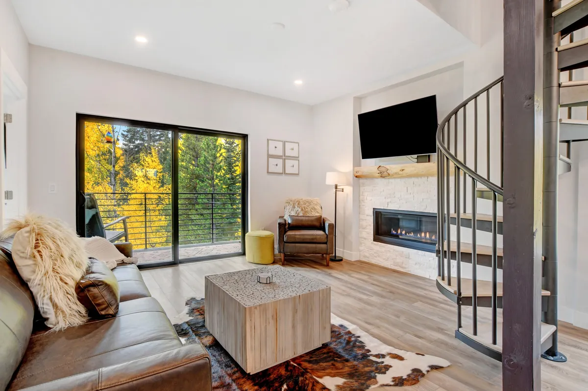This inviting living area features a modern gas fireplace, a comfortable seating arrangement, large glass doors leading to a patio, and a spiral staircase. The contemporary decor enhances the warmth, perfect for relaxing or socializing by the fire.