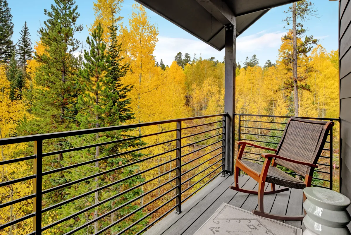 A tranquil balcony scene featuring a sturdy wooden railing, a classic rocking chair facing vibrant, multi-colored trees, and a cozy floor mat. The backdrop suggests either a golden autumn or lush spring, inviting relaxation and appreciation of the changing Colorado seasons.
