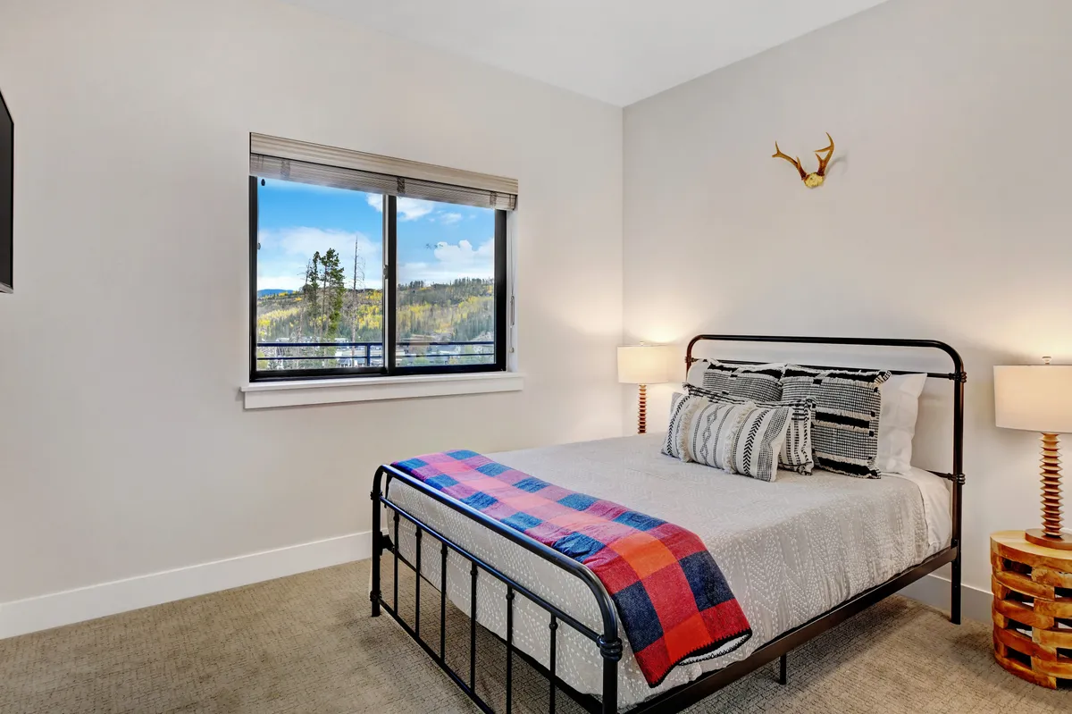 A tranquil bedroom featuring a comfortable bed with patterned bedding, a nightstand and lamp, decorative antlers, and a window framing scenic mountain views. The room feels restful, cozy, and ideal for unwinding.