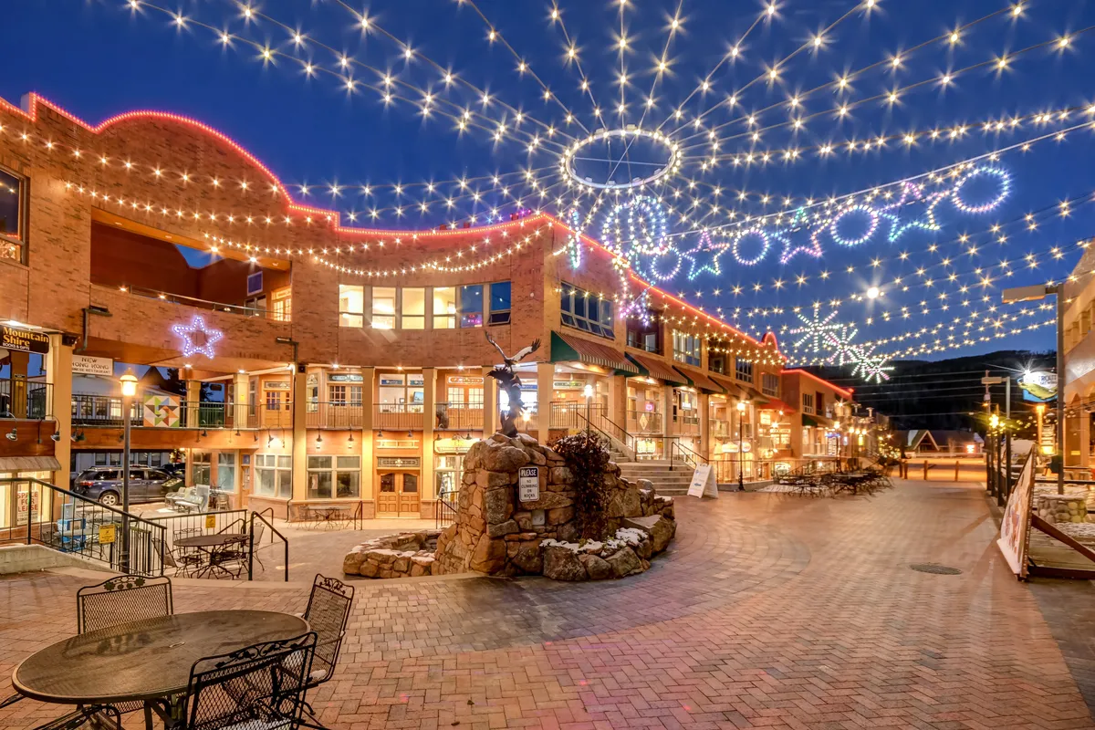 An exterior scene of downtown's festive ambiance, with decorative string lights, a central rock fountain, inviting outdoor seating, and vibrant storefronts. The atmosphere is joyful and bustling, ideal for shopping, dining, and community events just steps away.
