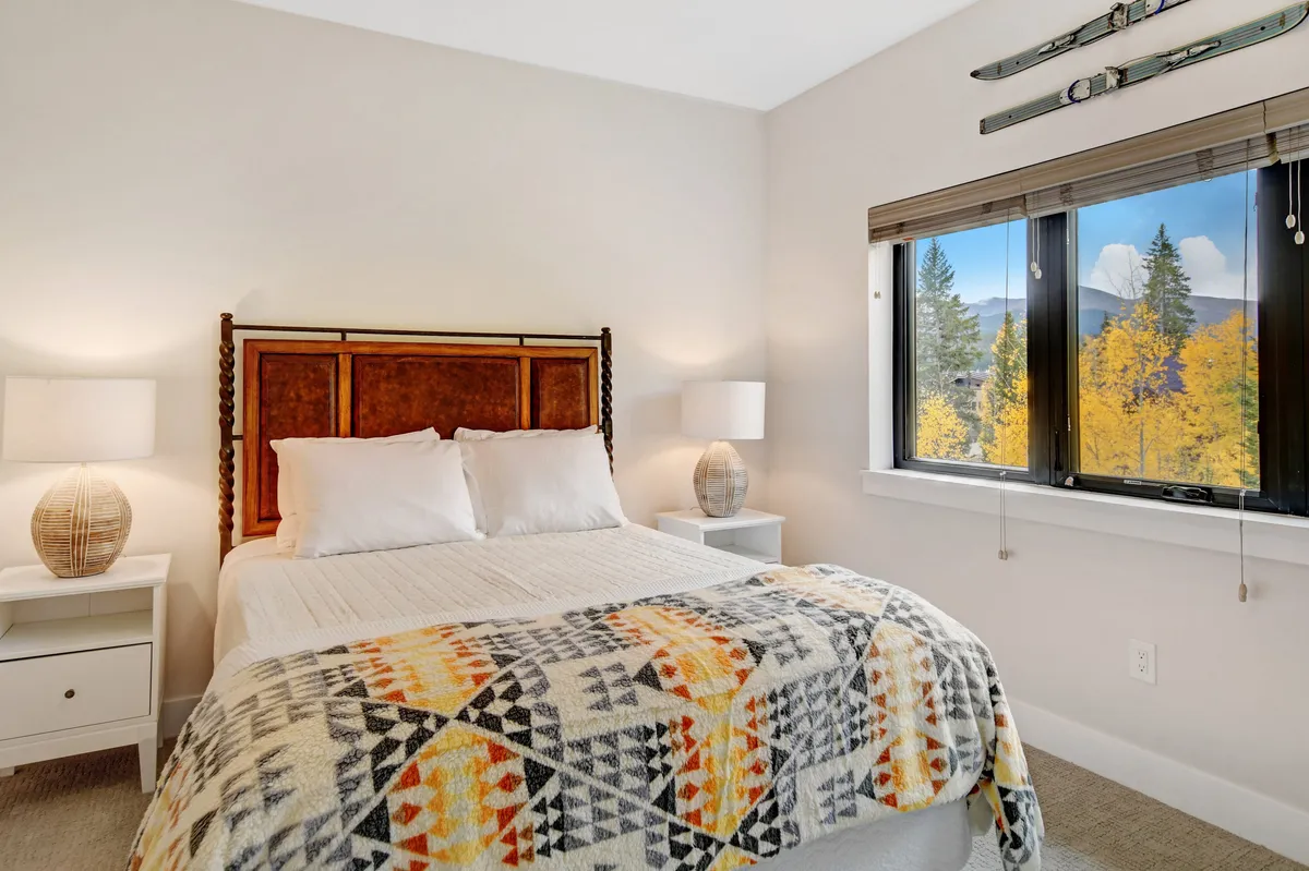 A serene bedroom featuring a plush bed with a decorative blanket, bedside tables with lamps, and a window framing mountain views. The tranquil mood invites relaxation and hints at premium bedding and restful comfort.